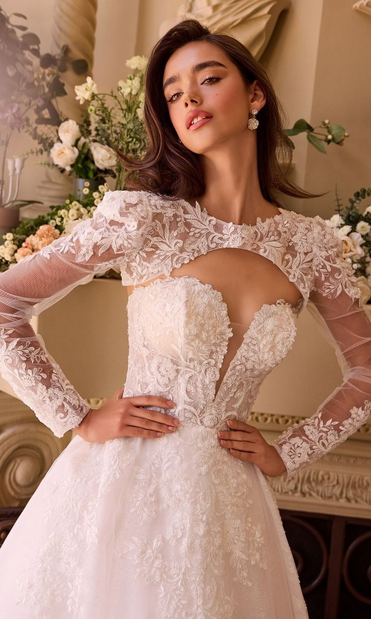 A woman wears the Andrea and Leo WL037 Long Wedding Dress, featuring an elegant lace bodice, long sheer sleeves with floral embroidery, and a romantic tulle skirt, standing indoors before a floral arrangement.